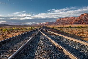 Railroad tracks converging in a desert landscape with red cliffs under a blue sky.