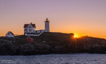 Lighthouse on rocky shore at sunset with vibrant sky.
