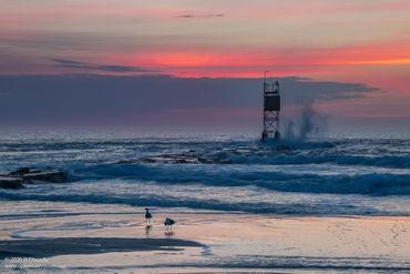 Sunset over ocean waves with a tower and birds on the shore.