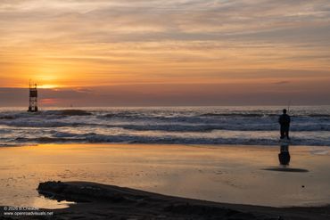 A person fishing on the beach at sunset with a lighthouse in the background.