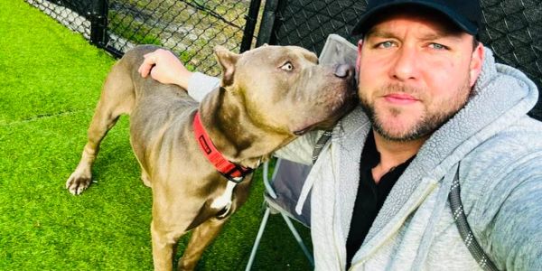 Man takes a selfie while a dog affectionately nuzzles his face.