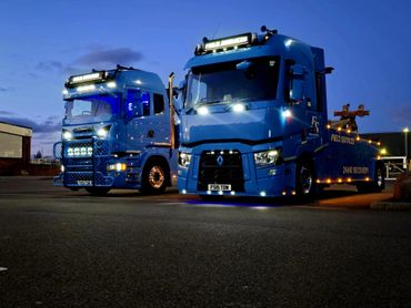Two illuminated blue trucks parked at dusk with lights on.