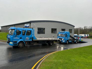 Two blue tow trucks parked near a gray industrial building on a wet road.