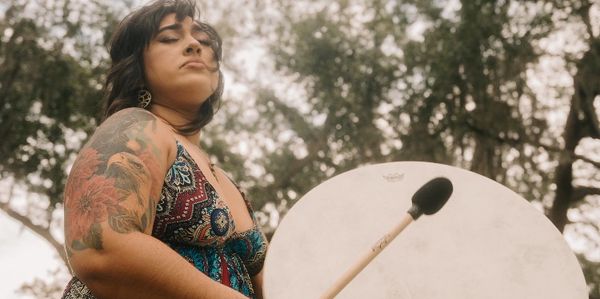Dark haired woman with her eyes closed playing large drum wearing burgundy dress