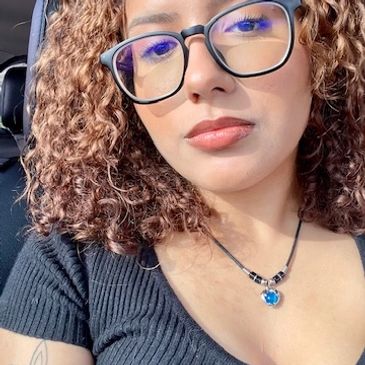 Woman with curly hair and glasses wearing a black top and blue pendant necklace.
