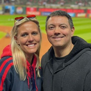 Smiling couple posing at a sports stadium with a green field in the background.