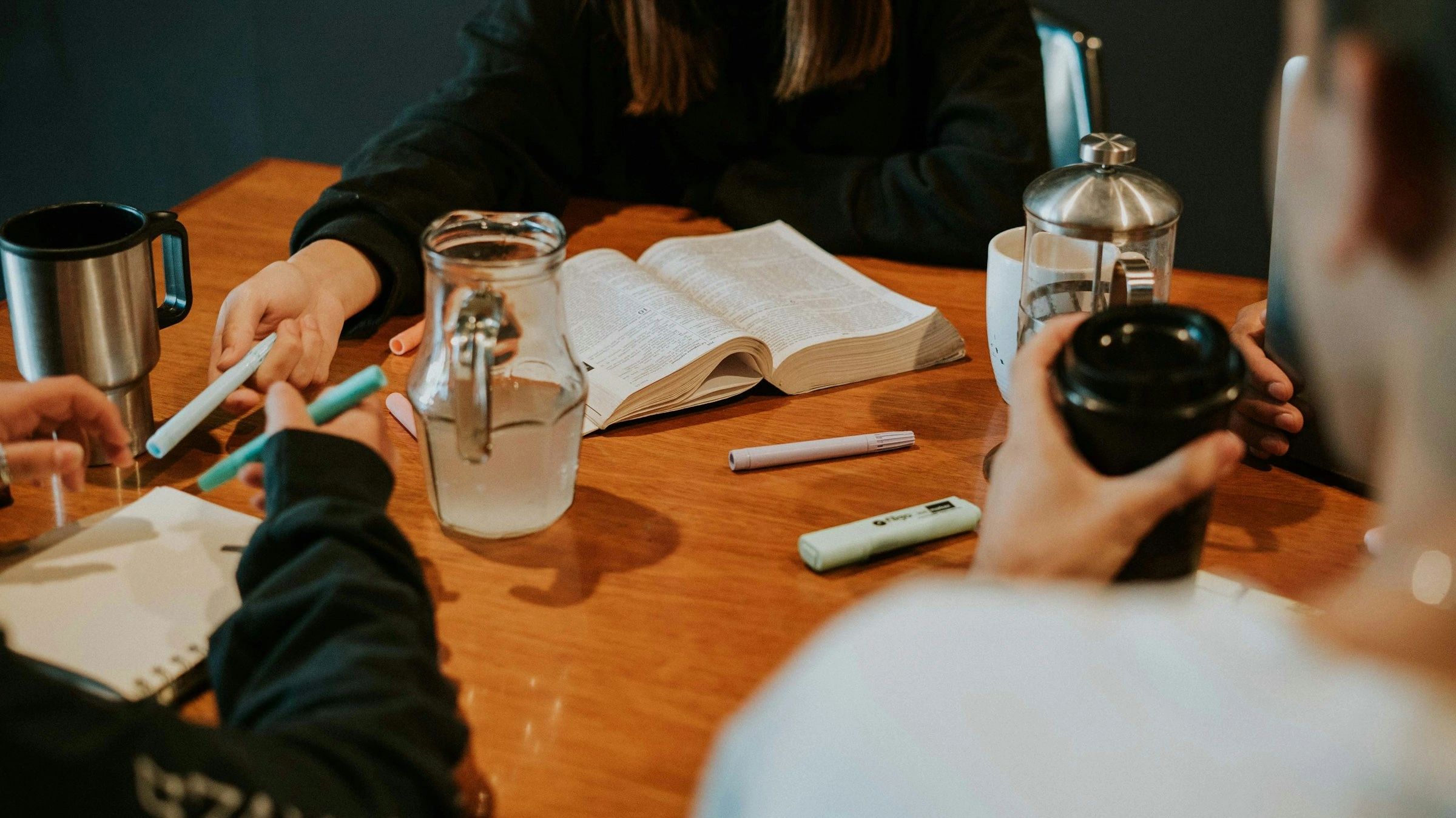 People studying together with books, markers, and drinks on a wooden table.