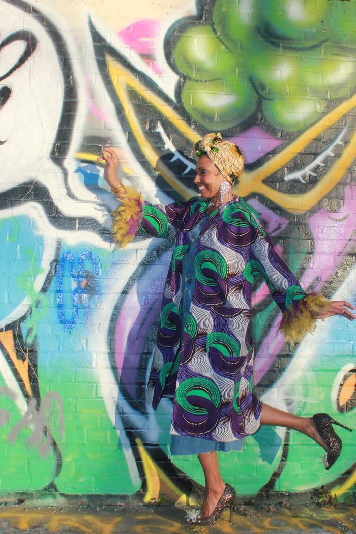 Woman running in front of a graffiti covered wall wearing a head wrap and jacket with feather trim.