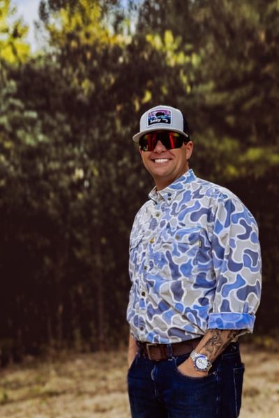 Man in camo shirt and cap smiling outdoors.