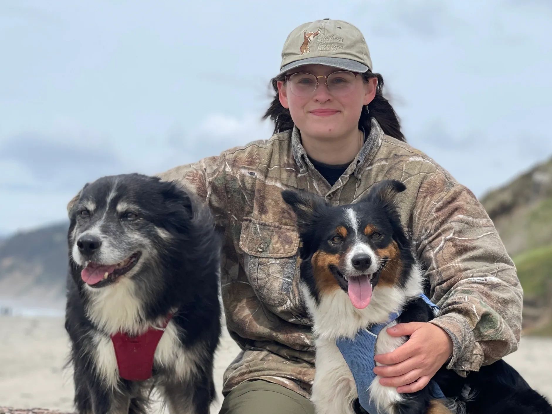 person in their 20s kneels on the beach with an arm around each of their dogs, both border collies