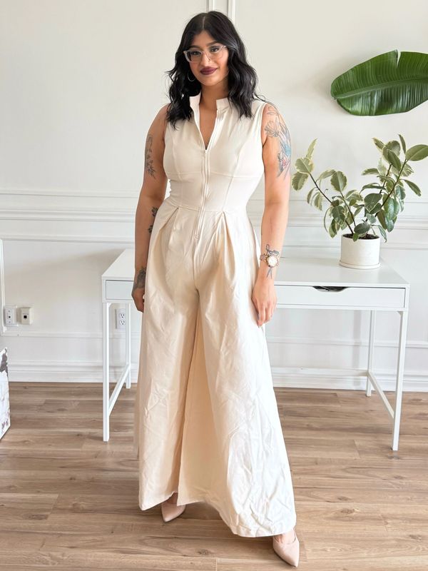 Woman in a cream sleeveless jumpsuit with tattoos, glasses, and dark hair, standing indoors.