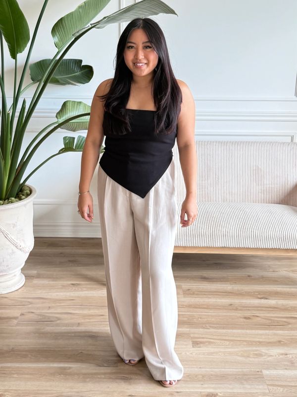 A woman stands indoors wearing a black top and beige pants, smiling at the camera.