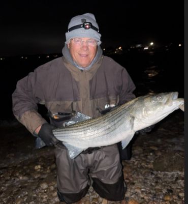 Man holding a large striped fish at night, dressed in fishing gear.