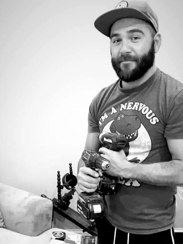 Man wearing a cap and dinosaur shirt holding two power drills indoors.