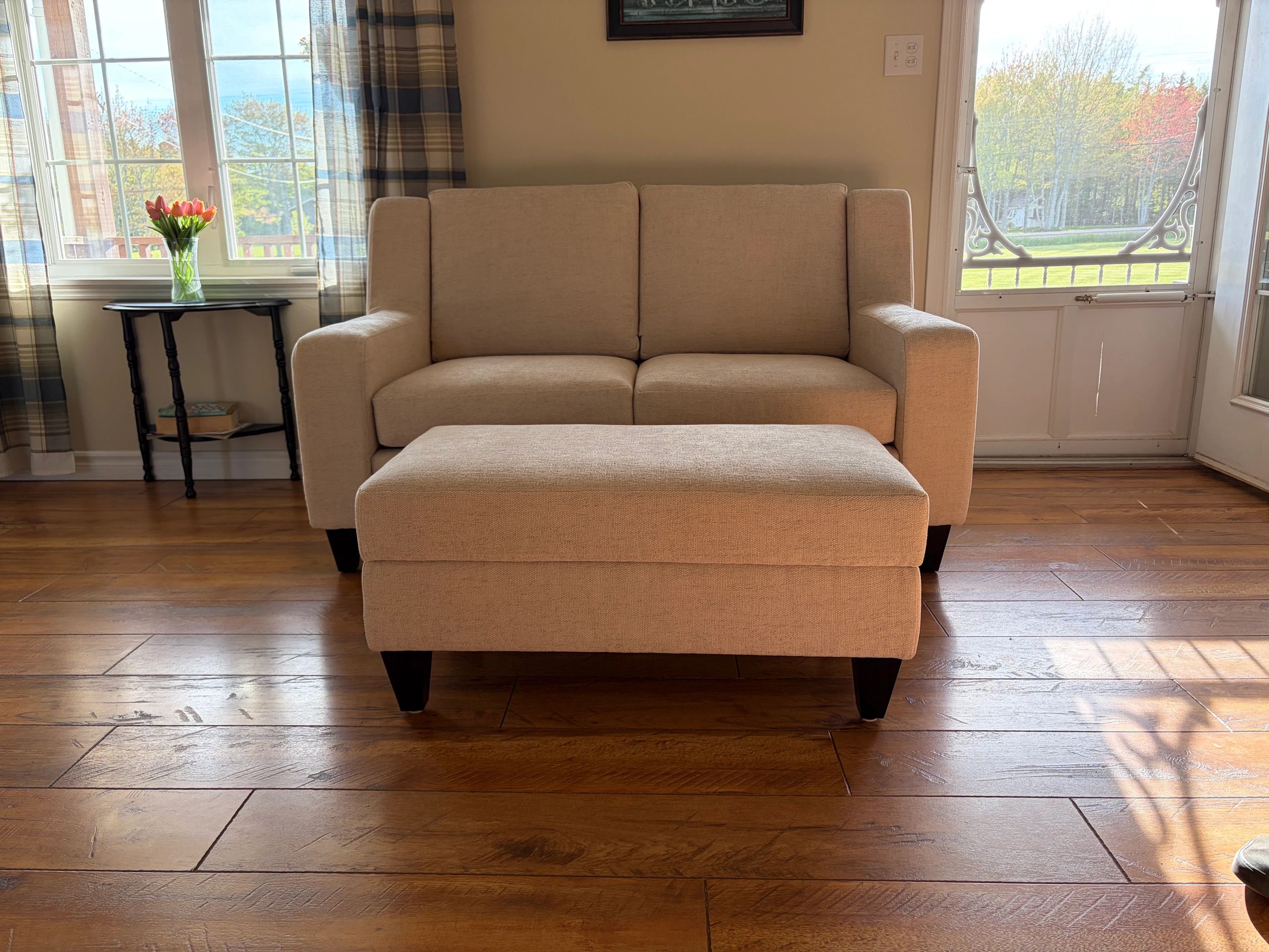Cozy beige loveseat and ottoman with sunlight streaming through windows.