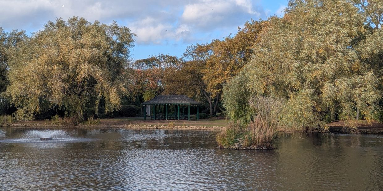 A peaceful park pond with fountain and gazebo surrounded by autumn trees.