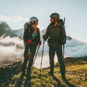 Two hikers with helmets and backpacks stand on a mountain trail at sunrise.