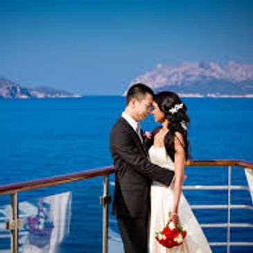 Bride and groom embrace on a yacht with ocean and mountains in the background.