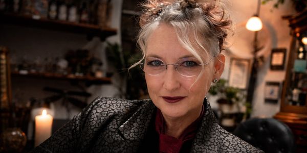 A confident woman with glasses and an intricate updo in a warm, atmospheric space, framed by rich tones, soft light, and an artist’s presence.