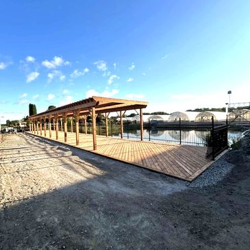A long wooden pergola near greenhouses and a pond under a clear blue sky.