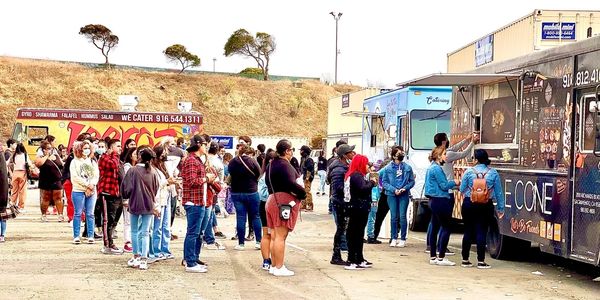 People waiting in line at food trucks outdoors on a clear day.
