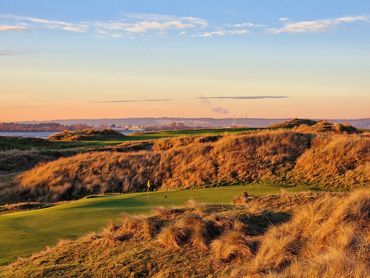 Sunlit golf course with rolling hills and distant industrial skyline at sunset.