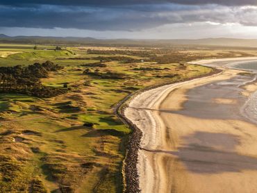 A coastal golf course next to a sandy beach under a dramatic sky.