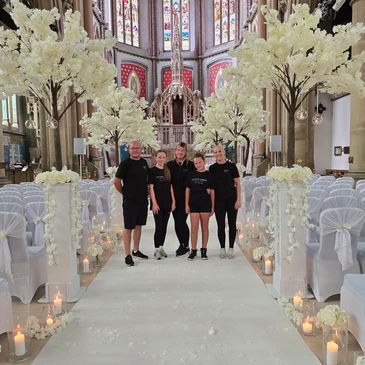 Five people posing in a beautifully decorated church aisle with white flowers and candles.