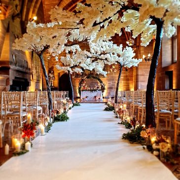 Elegant wedding aisle with white floral trees and candlelit decorations in a grand hall.