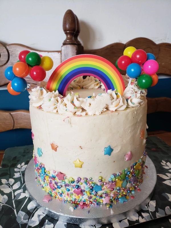 A white cake with a sprinkles border and a rainbow and balloons on top