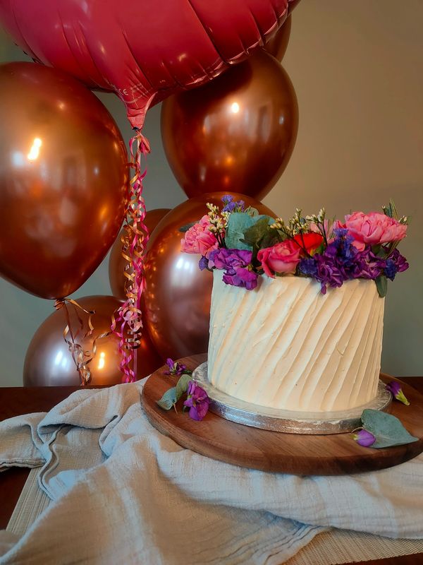 A white cake decorated with fresh flowers. Balloons in the background