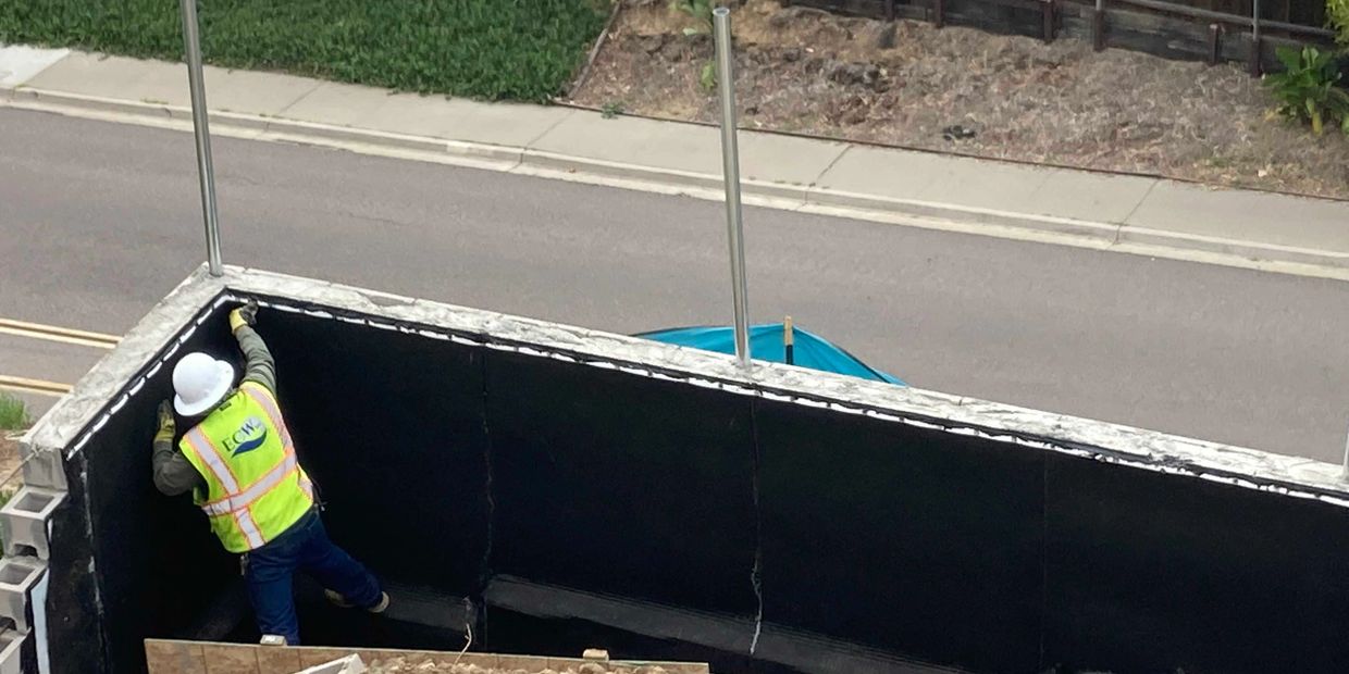 Construction worker applies material to a large black wall outdoors.