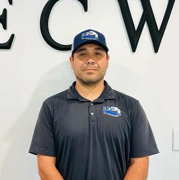 Man wearing a black polo shirt and matching cap stands against a white wall with large letters.