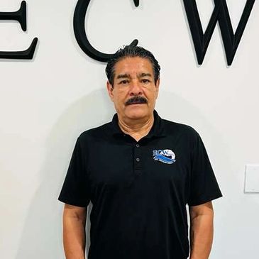 Man with mustache in black polo shirt standing against white wall.