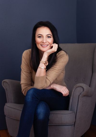 Smiling woman sitting comfortably in a gray armchair against a dark background.