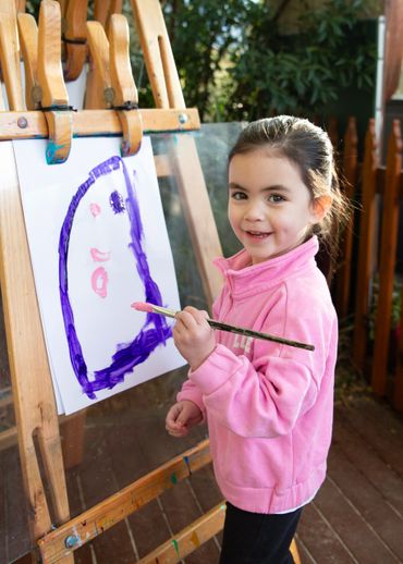 A young girl painting a colorful face on an easel outdoors.
