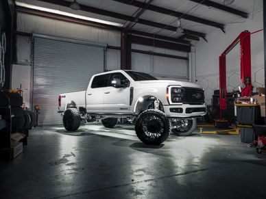 White lifted Ford truck inside a garage with large black wheels and suspension.