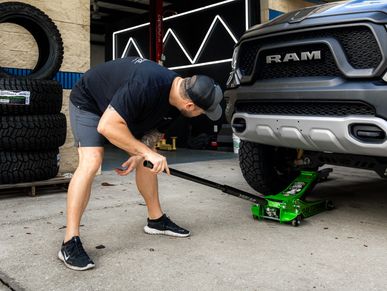 Man using a green jack to lift a RAM truck at a garage.