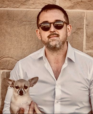 Man in sunglasses holding a small Chihuahua dog against a stone wall.