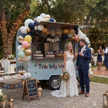 Newlyweds smile by decorated dessert cart at outdoor wedding.