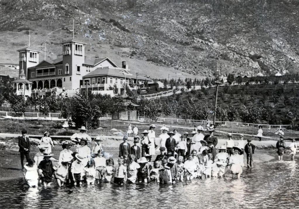A large group of people, mostly children, wading in shallow water near a grand building in a mountainous area.