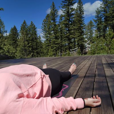 Person lying on a yoga mat on an outdoor deck amidst trees under a blue sky.