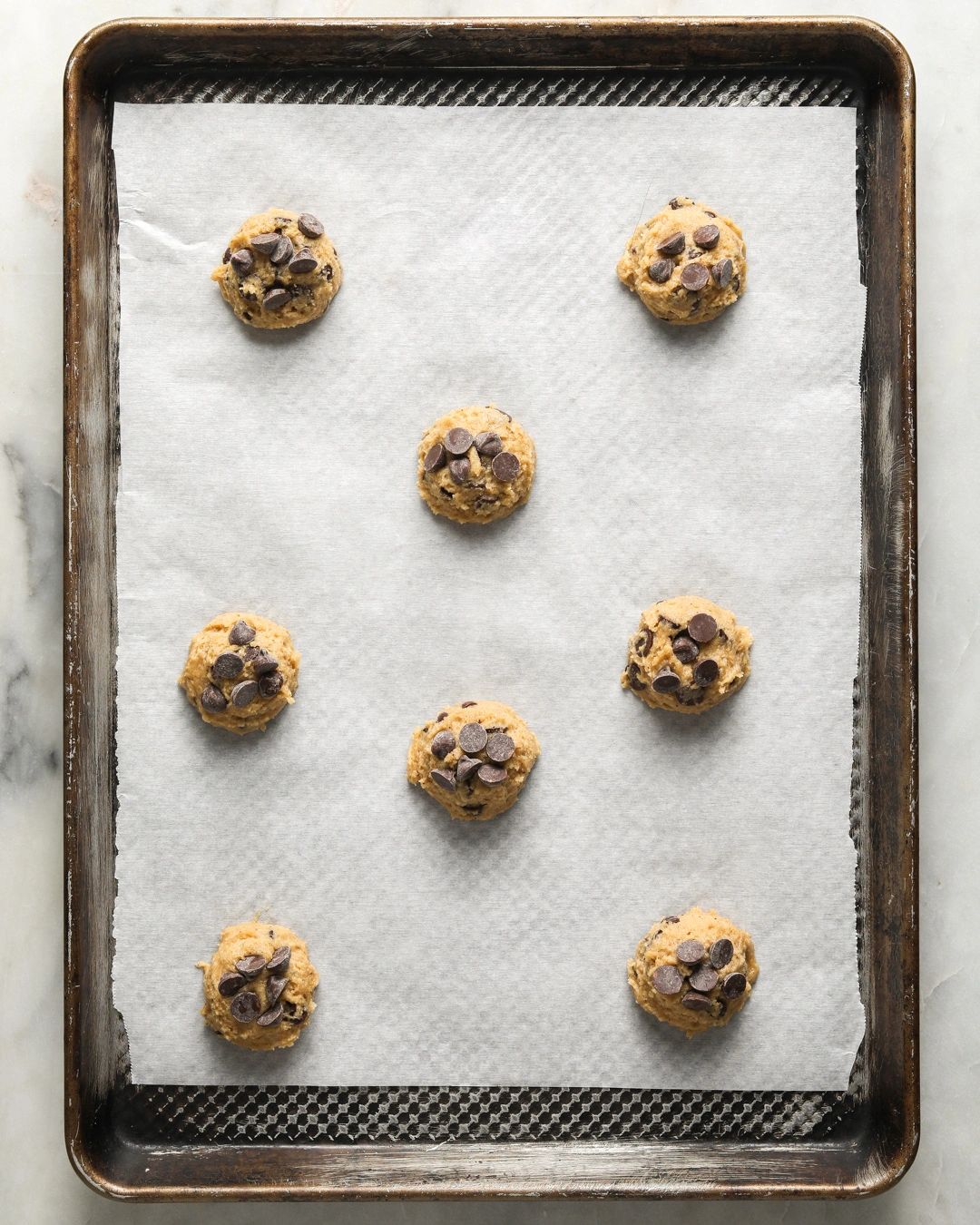 balls of brown butter chocolate chip cookie dough spread out on a half sheet baking tray, topped with extra chocolate chips