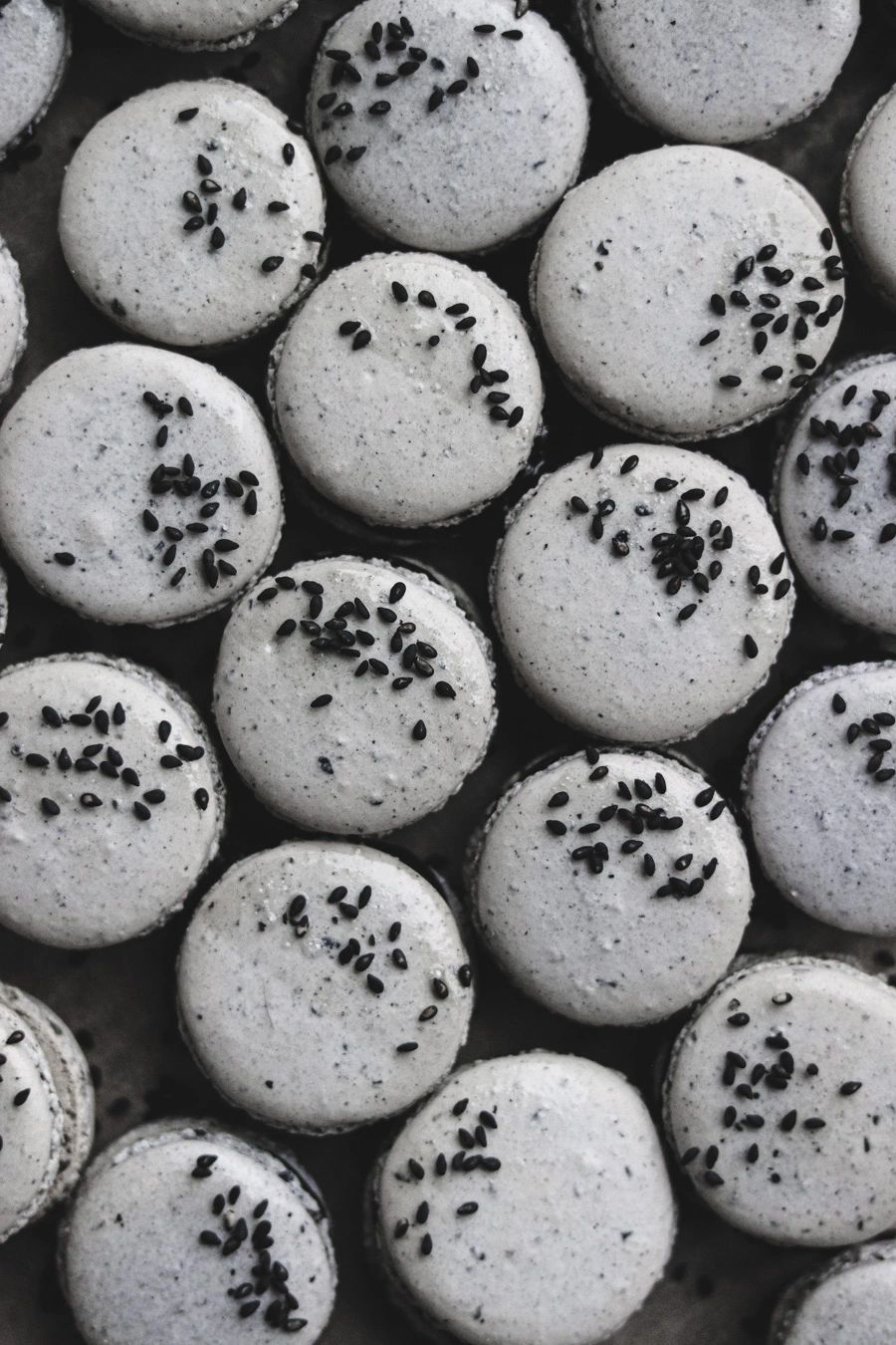 grey macaron shells topped with black sesame seeds crowded together on a baking sheet