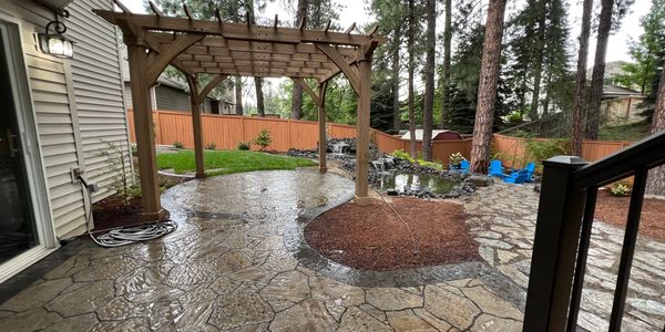 Backyard patio with pergola, stone flooring, and pond waterfall in a wooded setting.