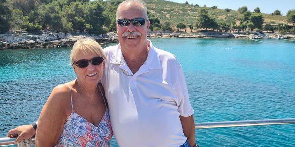 A smiling couple wearing sunglasses on a boat with turquoise water and greenery in the background.