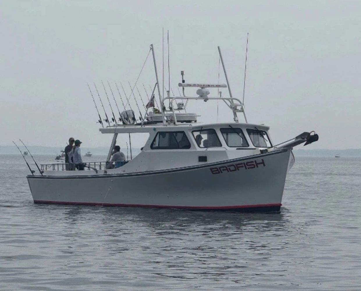 Fishing boat named BADFISH with people and multiple fishing rods on calm water.
