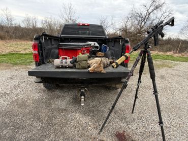Tripod-mounted rifle next to a pickup truck with gear and a dead animal in the bed.