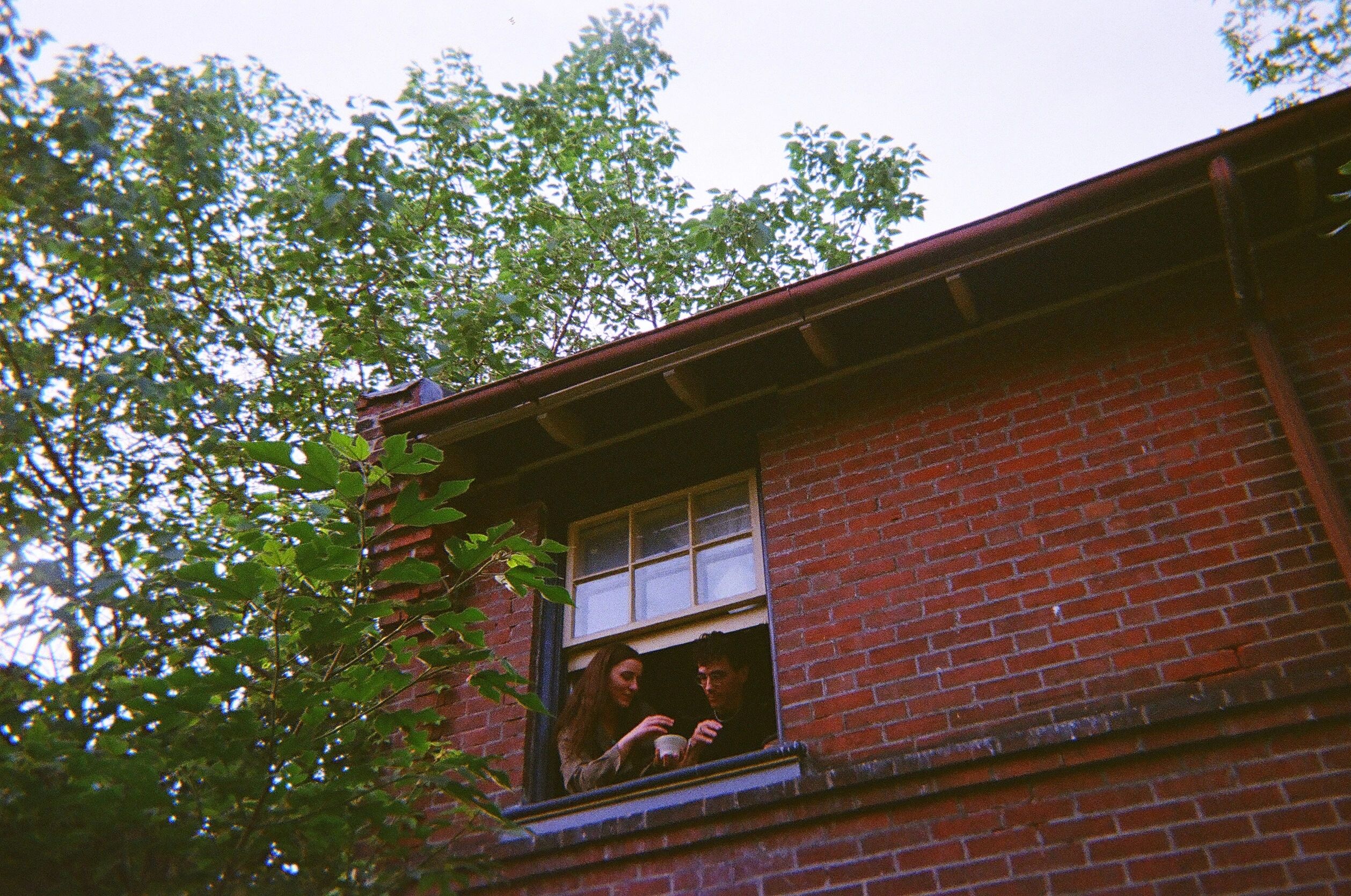 Two people chat by a window of a brick building surrounded by trees.