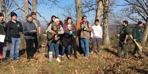 Group of people outdoors in a wooded area, some holding tools.